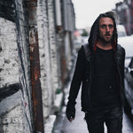 A man with long hair walks along a narrow street with a stone wall and parked cars. He wears the VENOM Men's Leather Hooded Vest Jacket with red lining from Kaya Movement, fingerless gloves, and distressed pants. The scene is dimly lit, creating a moody atmosphere.