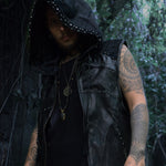 A person with tattoos on both arms stands in a forest, wearing the VENOM Men's Leather Hooded Vest Jacket by Kaya Movement. The person has a serious expression, and the black hood covers their head. They have multiple necklaces, including one with a pendant, and are surrounded by dense foliage.