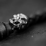 A close-up black and white image centers on a skull-shaped metal ornament, intricately detailed on a bracelet or strap. The background and the remainder of the strap are blurred, drawing sharp focus to the skull. This dark and dramatic tone evokes the essence of custom leather goods comparable to the edgy aesthetics of Kaya Movement's VENOM Men's Leather Hooded Vest Jacket with Red Lining.
