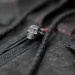 Close-up of a metal skull ring on a dark, textured surface with braided strings. Hints of red stitching in the background add contrast to the shadowy clothing, evoking the vibe of custom leather goods and a punk rock biker jacket, much like the VENOM Men's Leather Hooded Vest Jacket by Kaya Movement. The image has a gothic and mysterious feel.
