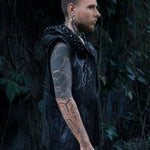 A side view of a person with tattoos on their arm and neck, wearing the Kaya Movement's VENOM Men's Leather Hooded Vest Jacket. They have piercings and gauges in their ear. The background features dense foliage and dark vegetation.