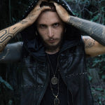 A tattooed person with piercings, wearing the VENOM Men's Leather Hooded Vest Jacket with Black Lining from Kaya Movement, stares intensely at the camera. They have various necklaces, including a pendant with a star symbol. They appear to be standing in a dimly lit forested area, surrounded by greenery.