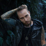 A person with tattoos on their arm and forehead, wearing a sleeveless black leather “VENOM Men's Leather Hooded Vest Jacket | Black Lining” by Kaya Movement, holds their hair up with one hand. The individual also has piercings and layered necklaces. They are standing among lush green foliage.
