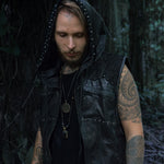 A man with tattoos on his arm and face stands in a forest. He wears a Kaya Movement VENOM Men's Leather Hooded Vest Jacket in black over a black shirt, with the hood partially covering his head. Sporting a pendant necklace, he appears to be looking down thoughtfully.