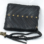 The AMONI Gold Stud Bi-Fold Flip Black Leather Wallet by Kaya Movement showcases a sophisticated chevron pattern and gold studs arranged in a line across the front. It features a braided leather strap with a brass clasp attached to the corner. This unisex designer wallet, highlighted against a plain white background, exemplifies exceptional craftsmanship and style.