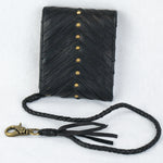 The AMONI Gold Stud Bi-Fold Flip Black Leather Wallet by Kaya Movement features a chevron pattern with small brass studs down the center, attached to a braided leather strap with a metal clasp. Tassels and a Labradorite stone accentuate the strap, all set against a plain white background.