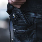 Close-up of a person's hand pulling an AMONI Black Stud Bi-Fold Flip Leather Wallet with Labradorite from the back pocket of their black jeans. This Kaya Movement wallet, featuring a textured surface and a decorative blue-stone button, complements the detailed stitching and ribbed fabric around the pocket area with its unisex designer appeal.