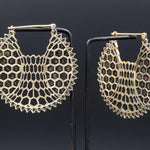The image features a pair of ILLUSION Oversized Cyborg Sci Fi Disc Earrings in Gold from Kaya Movement, showcasing an intricate honeycomb pattern design that evokes a post-apocalyptic aesthetic. They are elegantly displayed against a black background, hanging from thin metallic stands.