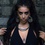 A person with long dark hair and intense makeup faces the camera. They are wearing a black halter top, post-apocalyptic jewelry, a necklace, and have multiple facial piercings. The ILLUSION Oversized Cyborg Sci Fi Disc Earrings in Gold by Kaya Movement add to the edgy look as they stand against a rugged brick wall backdrop, hands raised near their shoulders.