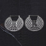 A pair of ILLUSION oversized cyborg sci-fi disc earrings in silver by Kaya Movement with an intricate honeycomb pattern on a dark textured background. These 14 gauge circular earrings feature delicate geometric designs extending from the center to the edges, adding sci-fi vibes to your look.