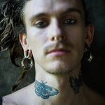 A person with long hair, adorned in the unisex PROMETHEUS Medium Biomechanical Hoop Earrings in Gold by Kaya Movement and a septum piercing, gazes ahead. They have intricate butterfly tattoos on their neck and upper chest. The background is dark and blurred, drawing focus to their facial features and accessories.