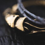 A close-up shot of the PROMETHEUS Small Cyborg Hoop Earrings in Gold by Kaya Movement, lying on a dark surface. The earrings showcase intricate details that evoke the feel of post-apocalyptic body jewelry, with visible texture and shadows enhancing their craftsmanship.