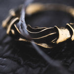 A close-up photo of the PROMETHEUS Small Cyborg Hoop Earrings in Gold by Kaya Movement, showcasing their intricate wavy detailing on a dark surface. A thin, blurred twig extends diagonally across the earrings, adding contrast to the textured background. The overall mood is moody and dramatic, resembling post-apocalyptic body jewelry.