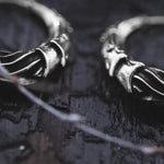 Close-up of the PROMETHEUS Small Cyborg Hoop Earrings, crafted from 925 silver by Kaya Movement, placed on a textured, dark surface. The earrings feature intricate wavy designs with subtle highlights and shadows emphasizing their detailed craftsmanship. A blurred element runs diagonally across the frame in the foreground.
