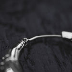 A close-up image of Kaya Movement's PROMETHEUS Small Cyborg Hoop Earrings in 925 silver, depicted against a dark, textured background. The photo zeroes in on a small segment of the curved metal, emphasizing its polished surface and intricate design details reminiscent of post-apocalyptic body jewelry.