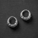 Displayed against a dark background, the PROMETHEUS Small Cyborg Hoop Earrings 925 Silver | 18 gauge by Kaya Movement feature intricate detailing resembling overlapping bands or layers, giving them a textured, almost post-apocalyptic body jewelry appearance.