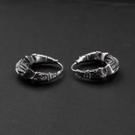 Displayed on a dark, flat surface is a pair of PROMETHEUS Small Cyborg Hoop Earrings by Kaya Movement, crafted from 925 silver. The intricate, circular design features embossed patterns with textured and raised sections, giving these 18-gauge sci-fi statement earrings a unique, handcrafted appearance.