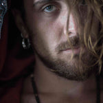 Close-up of a person with light blue eyes looking into the camera. The person has a beard and a mustache, and a strand of their light brown hair falls across their face. They are wearing PROMETHEUS Small Cyborg Hoop Earrings in 925 silver from Kaya Movement, along with post-apocalyptic body jewelry including a necklace. The background is blurred and dark.