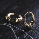 A close-up of two intricately designed gold hoop earrings on a dark textured surface. The PROMETHEUS Small Cyborg Hoop Earrings by Kaya Movement feature detailed designs that resemble skeletal fingers. Some thin, dried twigs lie nearby, adding to the moody, post-apocalyptic ambiance of the image.