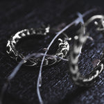 Close-up of two intricately designed Quetzalcoatl Oversized Snake Spine Hoop Earrings in Silver White Brass (14 gauge) by Kaya Movement on a dark, textured surface. The earrings, adorned with detailed patterns, are entwined with thin, dried vines, adding a mystical and vintage aura to the scene reminiscent of post-apocalyptic body jewelry. The lighting highlights the elegance of these captivating pieces.