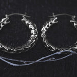 A pair of intricately designed Quetzalcoatl Oversized Snake Spine Hoop Earrings in Silver White Brass by Kaya Movement rests on a dark, textured surface. The earrings feature a detailed, overlapping leaf pattern, creating a bold and elegant look. Thin white strings lay nearby, adding to the contrast in this striking scene.
