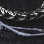 Close-up image of Kaya Movement's Quetzalcoatl Oversized Snake Spine Hoop Earrings in Silver White Brass, featuring intricate detailing that mimics small skulls or abstract shapes. The background is dark and slightly textured, with a thin, frosty white twig lying in front of the earrings, adding a striking contrast to the image—an exquisite piece that perfectly suits post-apocalyptic body jewelry.