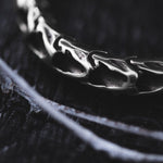 Close-up shot of Kaya Movement's Quetzalcoatl Oversized Snake Spine Hoop Earrings in Silver White Brass, resting on a textured black surface. The 14 gauge earrings feature a detailed snake spine design and are partially illuminated, evoking the look of post-apocalyptic body jewelry and creating a dramatic contrast with the background.