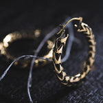 Close-up image of intricate QUETZALCOATL Oversized Snake Spine Hoop Earrings in Gold by Kaya Movement. They feature a textured, braided design and are placed on a dark, textured surface with frayed white string partially visible in the background. The soft lighting highlights the fine details of these sci-fi statement earrings.