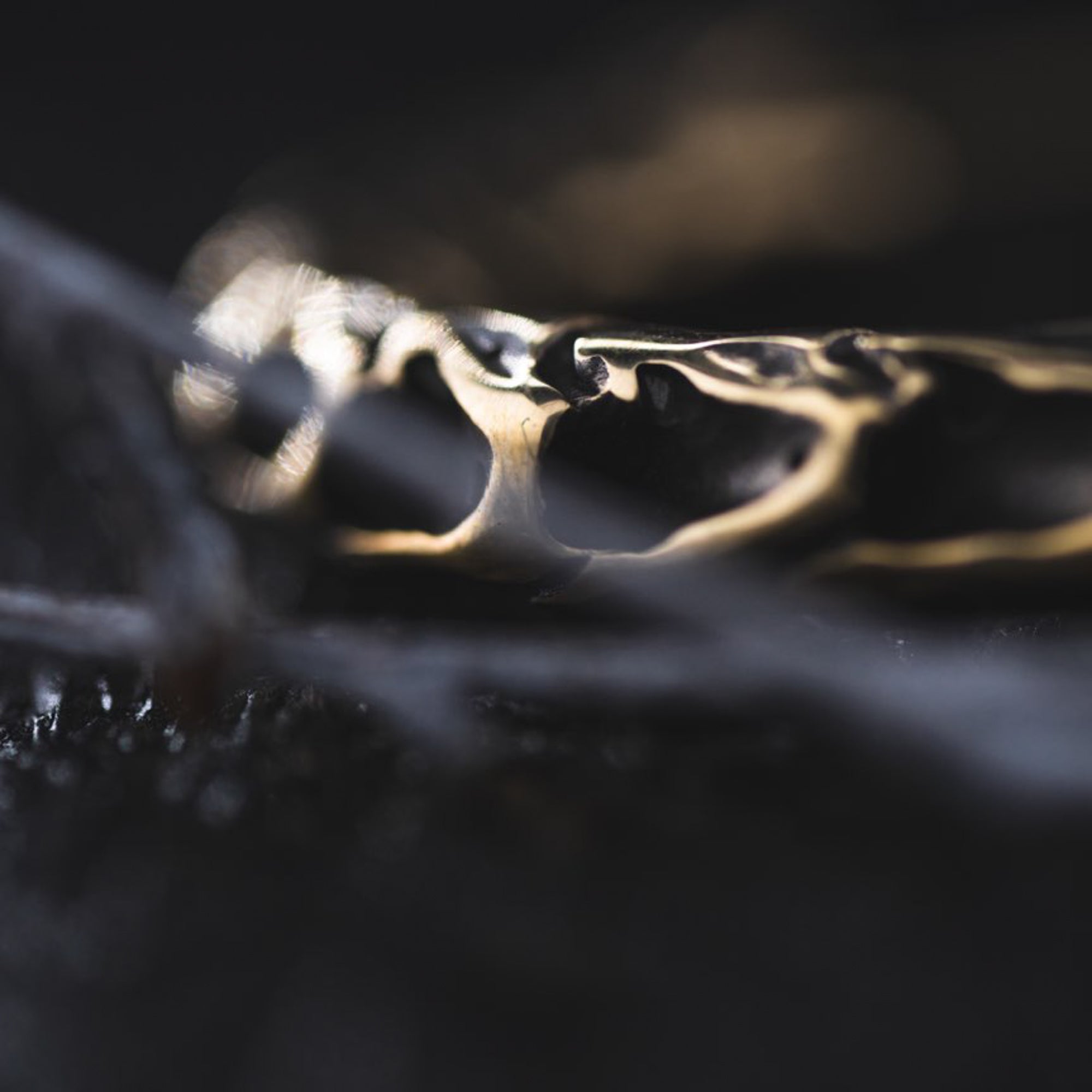 A close-up image of intricately detailed, gold-colored QUETZALCOATL Oversized Snake Spine Hoop Earrings in Gold by Kaya Movement on a dark surface. The focus is on the textured design of the earrings, which includes swirling patterns reminiscent of post-apocalyptic body jewelry. The background is blurred, emphasizing the earrings' craftsmanship and metallic sheen.