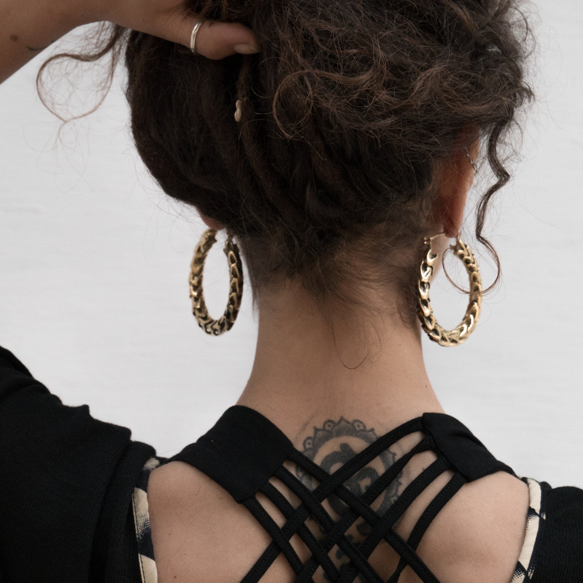 A person with curly brown hair styled in an updo is seen from behind. They are wearing QUETZALCOATL Oversized Snake Spine Hoop Earrings in Gold by Kaya Movement and a black top featuring intricate strappy details at the back. A tattoo is visible on the nape of their neck.