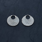 The SAYA Small Tribal Sayagata Endless Hoop Earrings in Silver by Kaya Movement, crafted from 925 sterling silver, are showcased against a dark textured background. These 18 gauge boho statement earrings feature an intricate maze-like geometric pattern and have a small loop at the top for attachment, embodying the essence of dark tribal body jewelry.