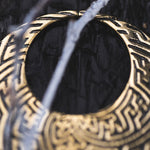 Close-up of the SAYAMOON Tribal Sayagata Seamless Hoop Disc Earring in Gold by Kaya Movement, with its 12-gauge intricate patterns, hanging on what appears to be a wire or branch. The dark background creates a striking contrast with the boho statement earring's details. The piece is partially obscured by out-of-focus elements in the foreground.