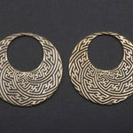 Lying against a dark background is a striking pair of SAYAMOON Tribal Sayagata Seamless Hoop Disc Earrings in Gold by Kaya Movement. These 12 gauge earrings feature intricate, maze-like patterns with geometric lines and shapes arranged in a symmetrical, circular layout, making them an eye-catching boho statement piece.