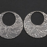The SAYAMOON Tribal Sayagata Hoop Disc Earrings in 925 Silver by Kaya Movement feature large, circular Sterling silver earrings with intricate, geometric patterns etched into the metal on a solid black background. These detailed and textured designs are reminiscent of tribal body jewelry. They are constructed in a 12 gauge size.