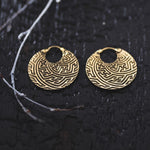 A close-up of the SAYA Small Tribal Sayagata Disc Hoop Earrings in Gold by Kaya Movement, showcasing their intricate geometric maze patterns reminiscent of traditional Sayagata. The 18-gauge boho statement jewelry is displayed on a black textured surface, partially bordered by small branch twigs.