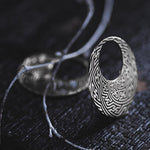 A close-up of the SAYAMOON Tribal Sayagata Hoop Disc Earrings in 925 Silver by Kaya Movement, featuring an intricately designed maze-like pattern, sits on a dark surface. Nearby, a thin, twisted branch lies partially blurred in the background, adding a natural element to this elegant piece of tribal body jewelry.