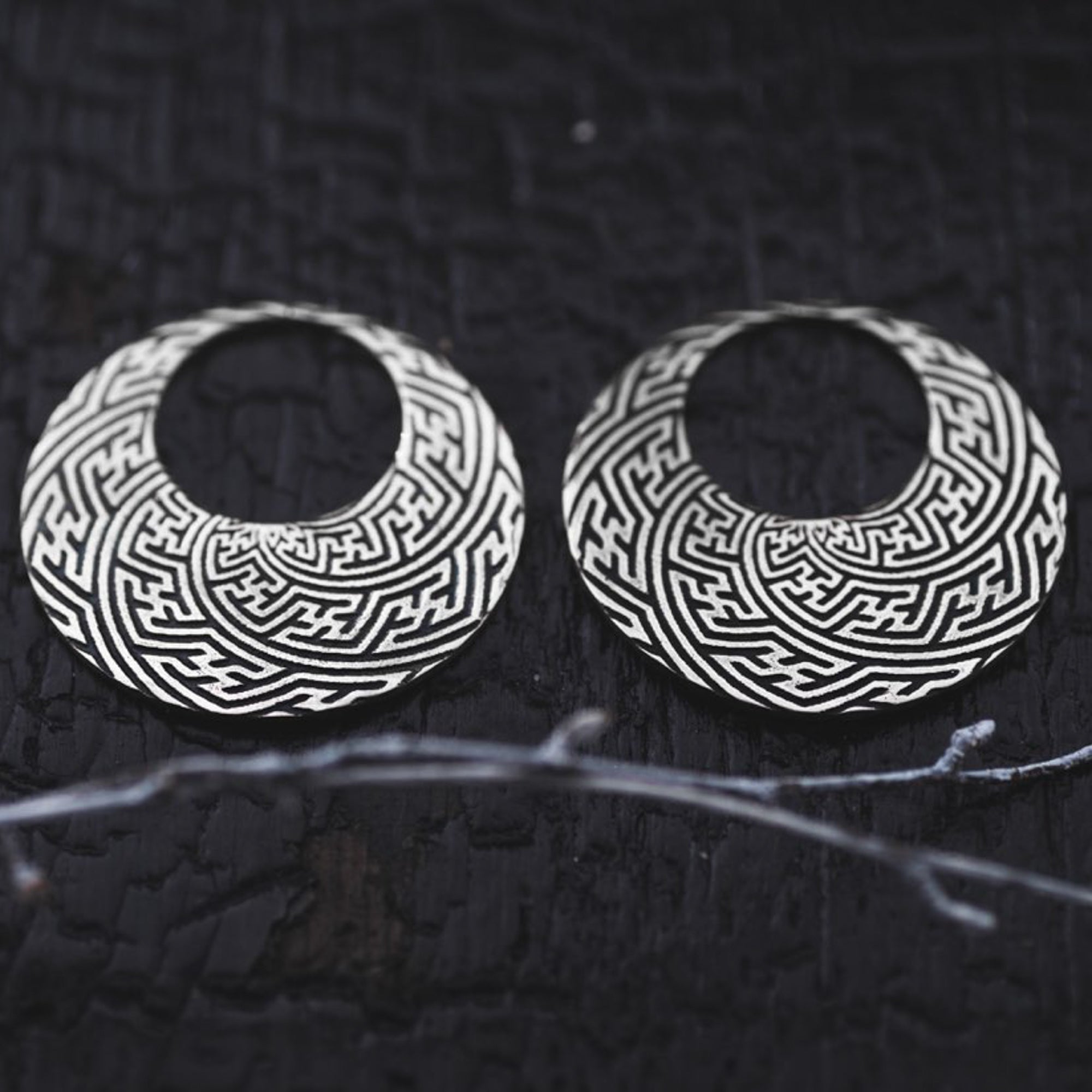 The SAYAMOON Tribal Sayagata Hoop Disc Earrings in 925 Silver by Kaya Movement feature a pair of round, Sterling silver earrings with intricate, maze-like patterns etched into the surface. They are beautifully displayed on a dark textured background, with a delicate branch in the foreground for added contrast.