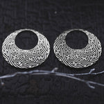 The SAYAMOON Tribal Sayagata Hoop Disc Earrings in 925 Silver by Kaya Movement feature a pair of round, Sterling silver earrings with intricate, maze-like patterns etched into the surface. They are beautifully displayed on a dark textured background, with a delicate branch in the foreground for added contrast.