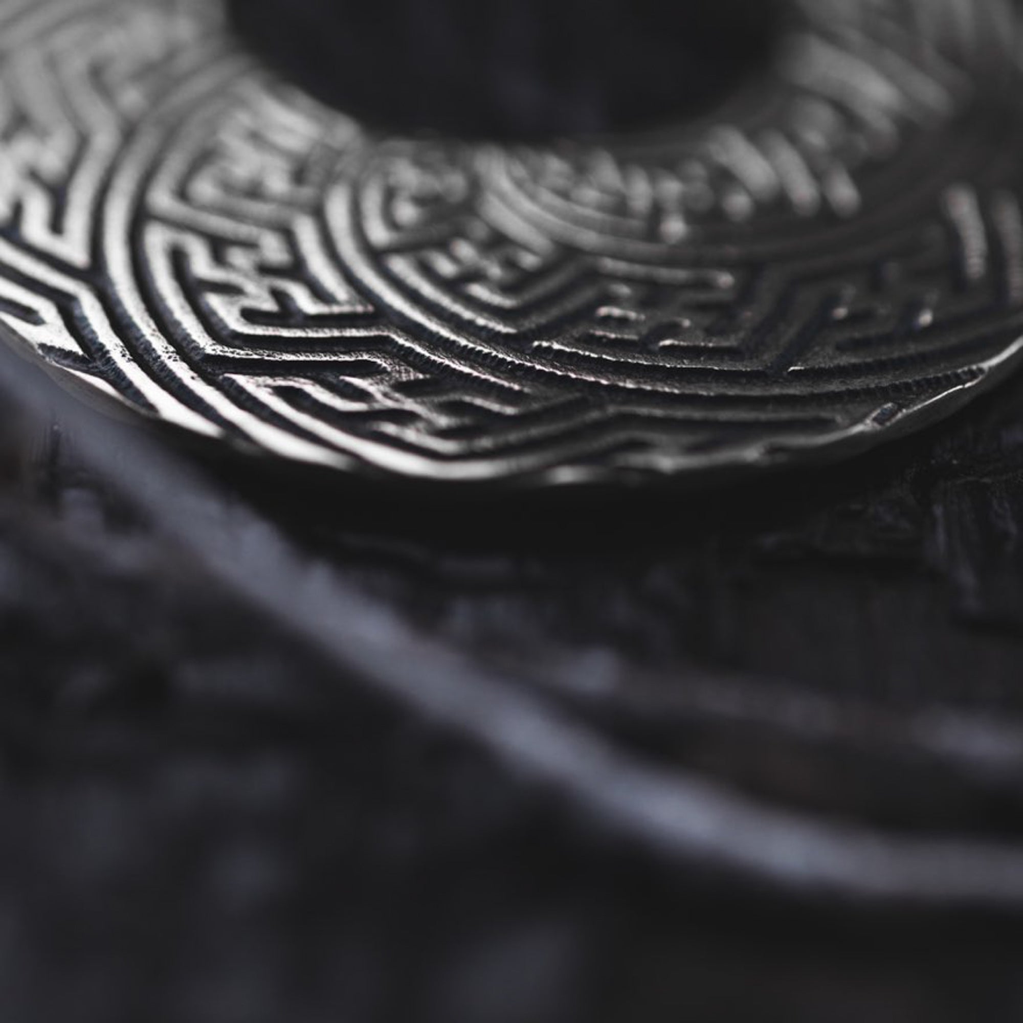 Close-up of tribal-inspired SAYAMOON Tribal Sayagata Hoop Disc Earrings in 925 Silver by Kaya Movement, featuring intricate maze-like engravings. The pendant earrings are displayed with a dark string slightly out of focus, against a dark background that highlights the detailed texture.