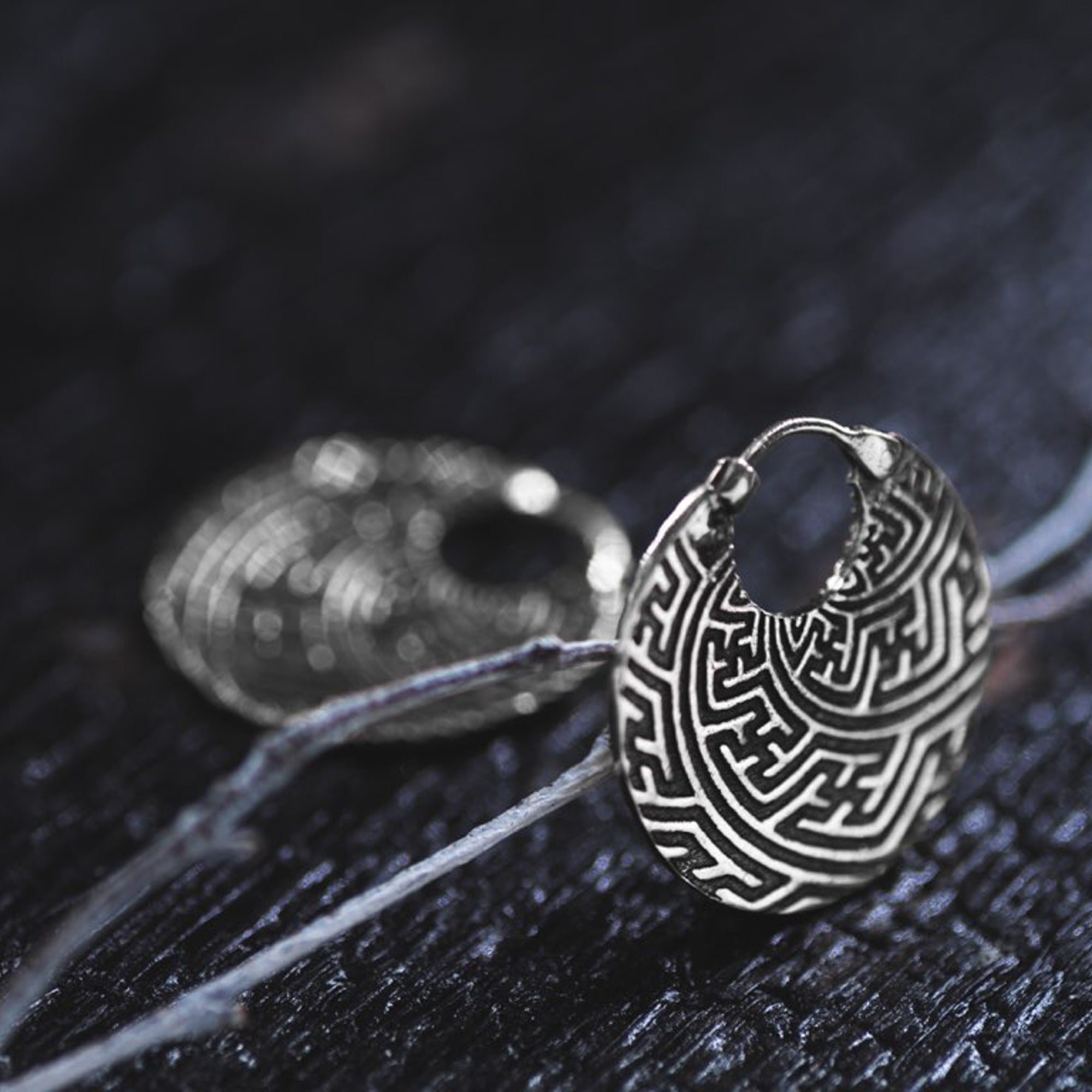 A close-up of the Kaya Movement's SAYA Small Tribal Sayagata Endless Hoop Earrings in 925 sterling silver, showcasing intricate maze-like patterns. The boho statement earrings are placed on a dark, textured surface, with one earring sharply focused in the foreground and the other artfully blurred in the background.