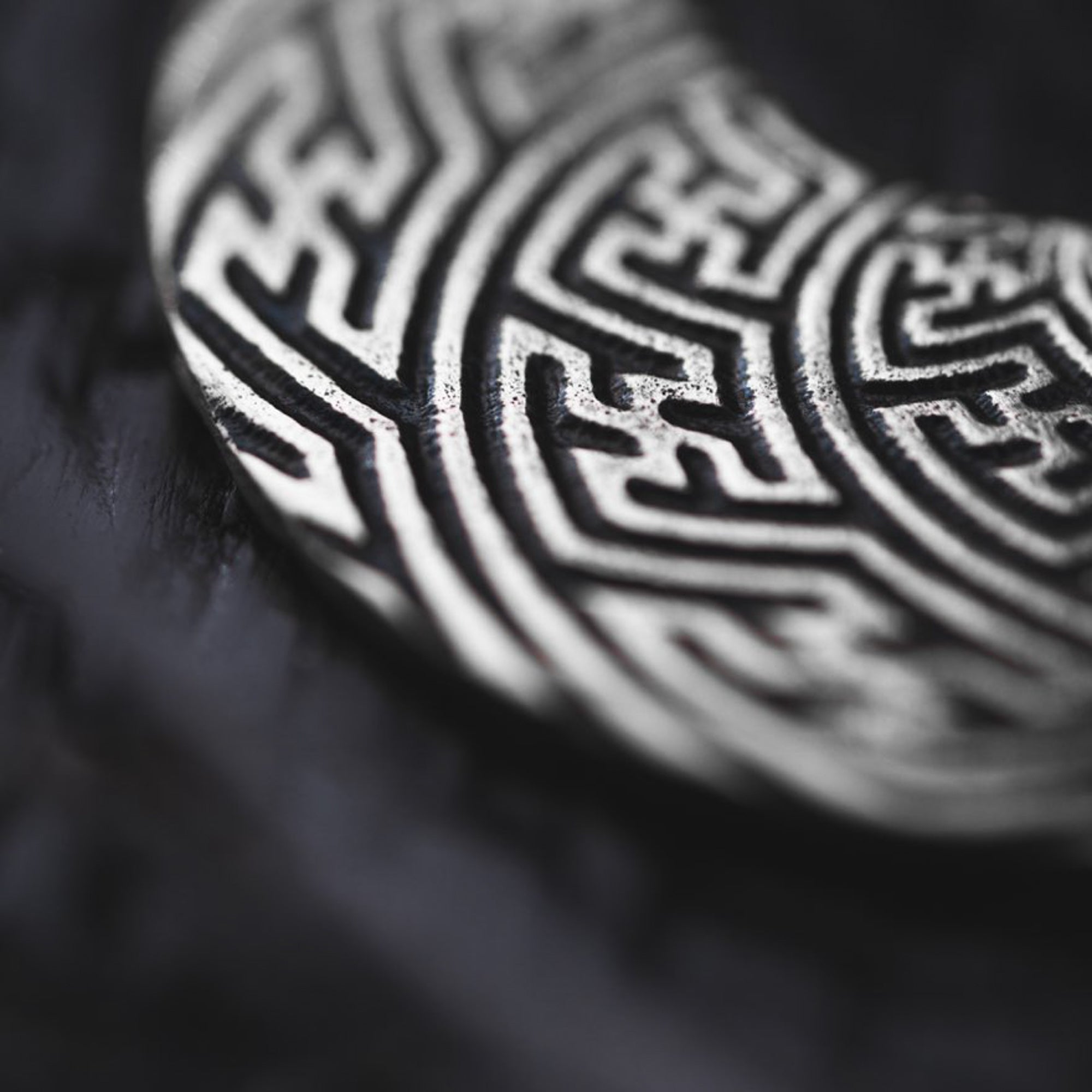 Close-up of the SAYAMOON Tribal Sayagata Hoop Disc Earrings in 925 Silver by Kaya Movement, showcasing their intricate, maze-like patterns on a metallic circular surface. The design is set against a blurred, dark background, highlighting the texture and detail of the engraving.