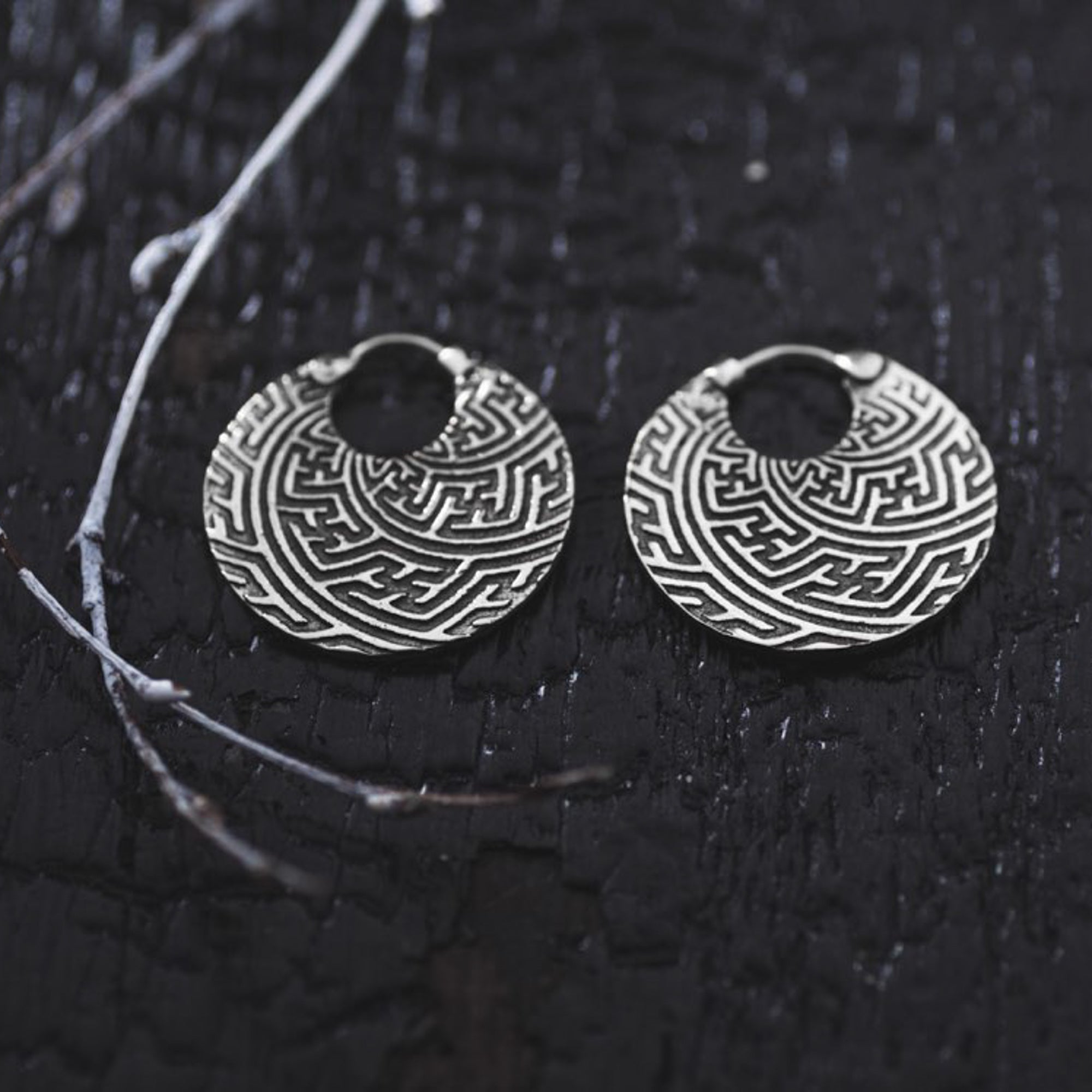 A pair of Kaya Movement's SAYA Small Tribal Sayagata Endless Hoop Earrings in 925 sterling silver, exquisitely designed with intricate maze patterns, are laid on a dark, textured surface. A small, dry twig with delicate branches is positioned diagonally across the background, adding contrast to these boho statement earrings of 18 gauge.