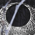 A close-up image of a pair of intricately designed SAYAMOON Tribal Sayagata Hoop Disc Earrings in 925 Silver by Kaya Movement, featuring geometric patterns. The earrings, reminiscent of tribal body jewelry, hang among blurred, translucent branches or wires with a dark, textured background that highlights the fine details of the metalwork and their 12 gauge thickness.