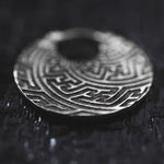 Close-up of a SAYA Tribal Sayagata Endless Hoop Earring in silver, lying flat on a dark, textured surface. The 18 gauge earring from Kaya Movement, reminiscent of dark tribal body jewelry, features intricate embossed patterns with interlocking geometric shapes, creating a visually striking labyrinth-like design. There's a small hole at the center of the earring.