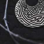 Close-up of the SAYA Small Tribal Sayagata Endless Hoop Earrings in Silver, showcasing their intricate maze-like engravings, resting on a dark, textured surface. A slender, dry branch is partially visible, intersecting the hoop earring. The image exudes a dramatic, high-contrast feel reminiscent of dark tribal body jewelry by Kaya Movement.
