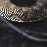 Close-up image of a dark, intricately patterned ring with a circular design. The ring features detailed geometric and maze-like engravings, similar to the Kaya Movement's SAYAMOON Tribal Sayagata Seamless Hoop Disc Earrings in Gold (12 gauge), with highlights and shadows accentuating the textures. The background is blurred, focusing attention on the ring's design.