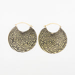 A pair of SAYAGATA Oversized Tribal Hoop Disc Earrings in Gold from Kaya Movement, featuring a round, intricately designed labyrinth-like pattern in gold and black. These boho statement earrings have a slightly domed shape and are secured with a simple gold clasp. They are displayed against a plain white background.