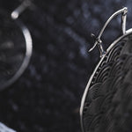 Close-up image of two intricately designed SEIGAIHA Oversized Japanese Geometry Disc Earrings in Silver by Kaya Movement, showcasing wave patterns on a dark, textured background. One earring is prominently in focus, revealing the fine details of the waves, while the other is blurred in the background.