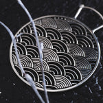 A SEIGAIHA Oversized Japanese Geometry Disc Earring in Silver from Kaya Movement, with an intricate pattern of semi-circular, wave-like designs, rests on a textured dark surface. Out-of-focus branches cross in front of the earring, adding depth and contrast to the image, reminiscent of boho statement pieces that capture a sense of natural elegance.