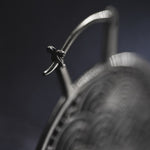 Close-up of the metal clasp on a decorative object, likely a piece of jewelry or adornment such as the SEIGAIHA Oversized Japanese Geometry Disc Earrings in Silver by Kaya Movement. The background is dark and out of focus, highlighting the intricate details and texture of the 16 gauge metalwork.
